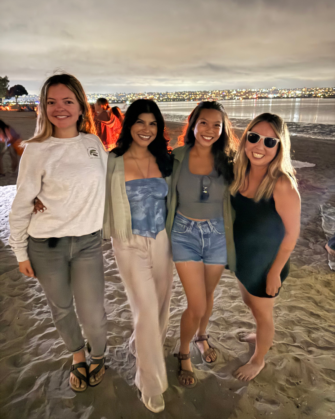 Four women standing on a sandy beach at night, smiling, with city lights reflecting on the water in the background.