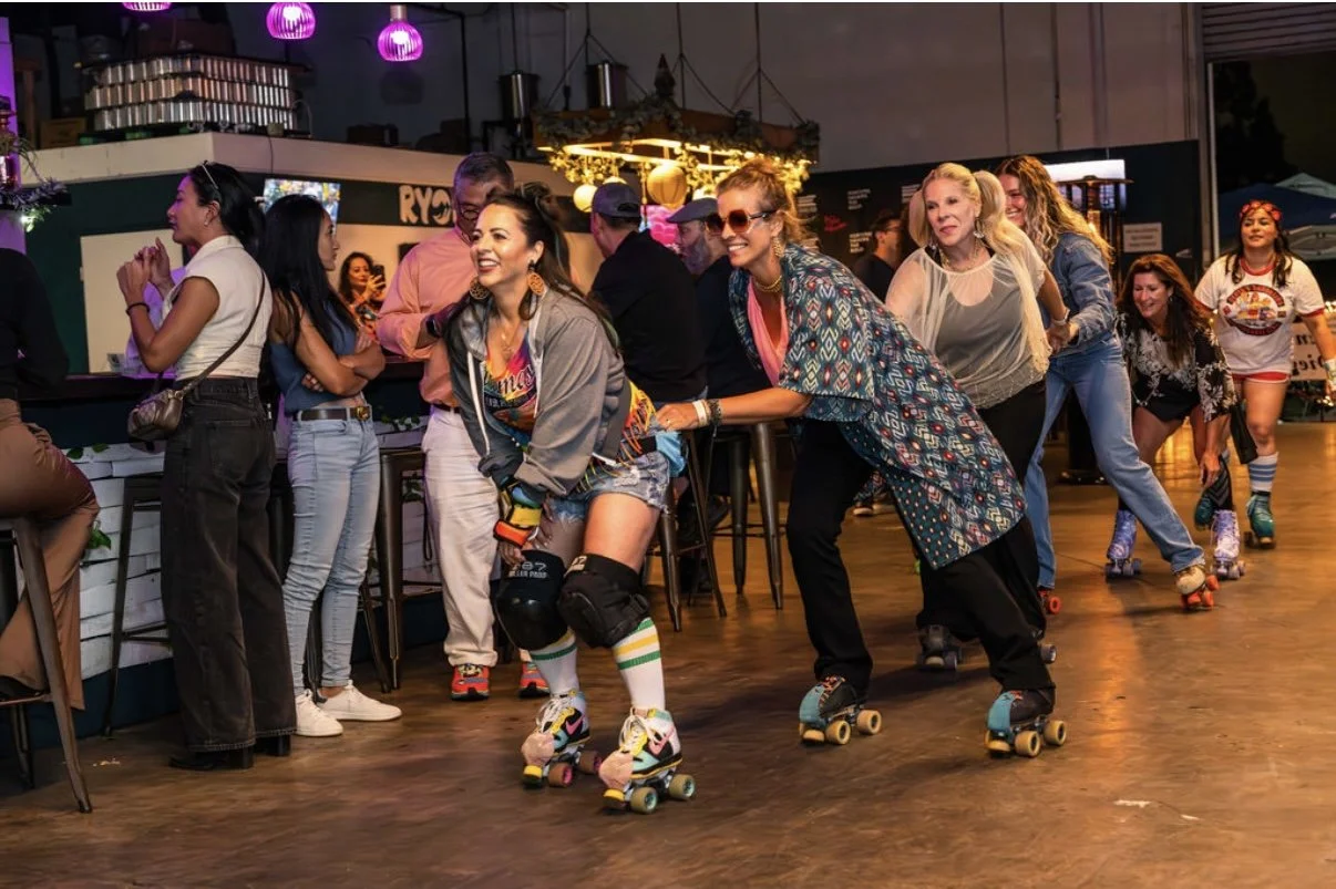 Group of people roller skating indoors at a lively retro-themed event, with participants laughing and forming a line while wearing colorful outfits in a vibrant social setting.