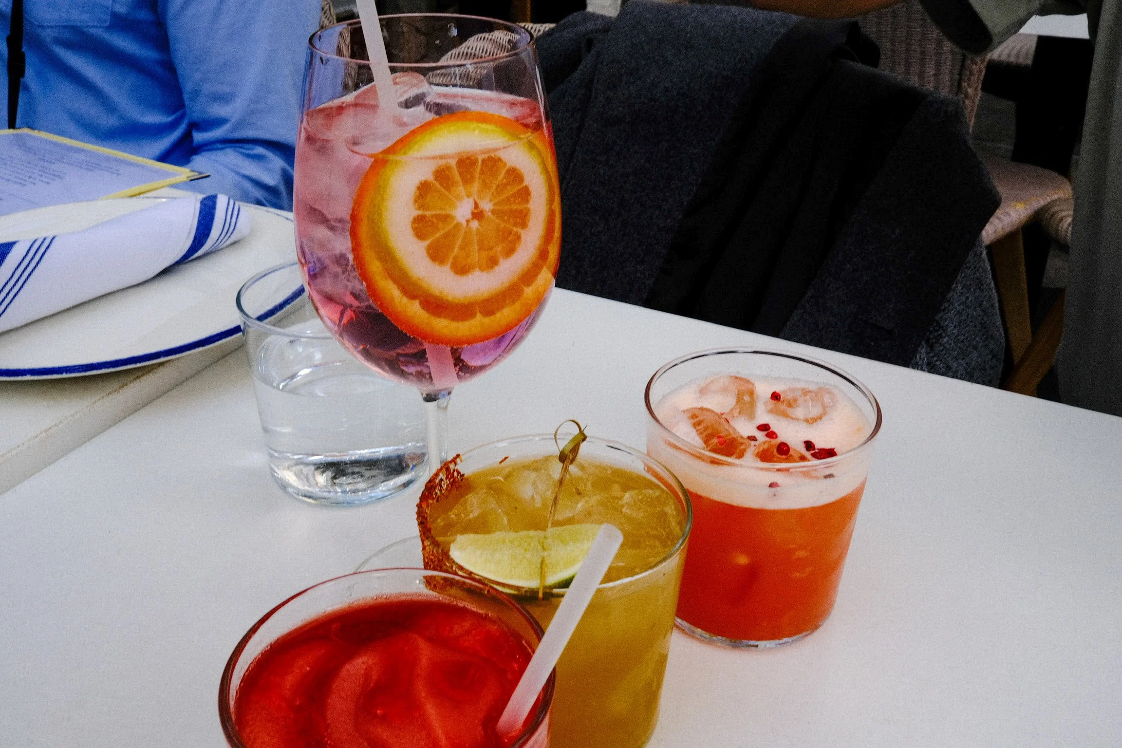 Close-up of vibrant, colorful cocktails on a table with a glass of water in the background. The drinks include a pink drink with orange slices, a yellow drink with a lime wedge, and a red drink with ice, creating a refreshing and inviting atmosphere.