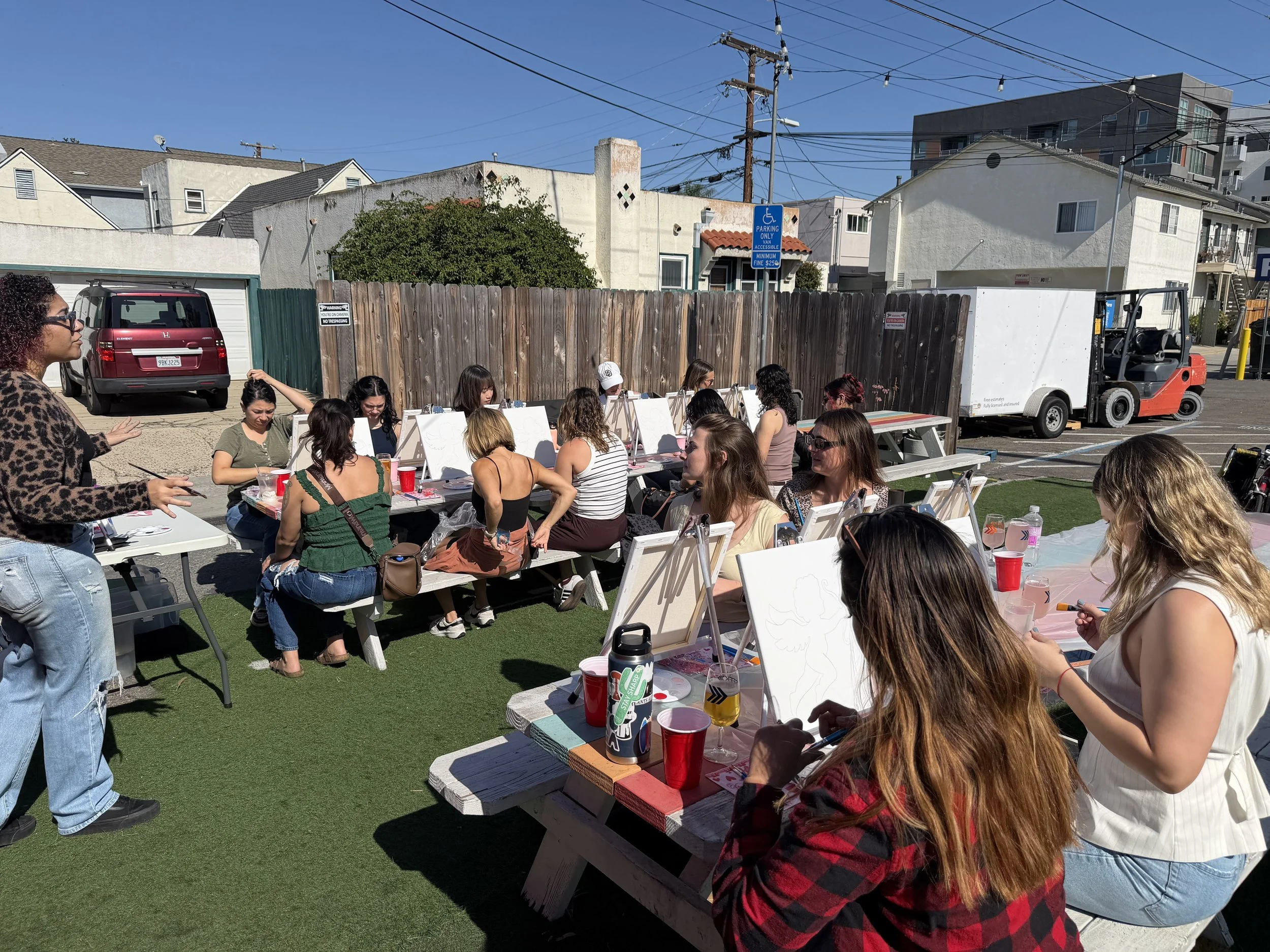 LetsGoGirls San Diego members enjoying a Paint & Sip event outdoors, painting on canvases at picnic tables while socializing, creating a fun atmosphere for friendship, creativity, and community connection.