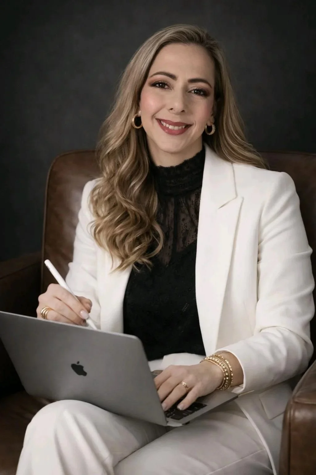 Carlota Alvarado sentada en un sillón de cuero, con traje blanco y blusa negra, trabajando en una laptop con lápiz digital y una expresión cálida y profesional.