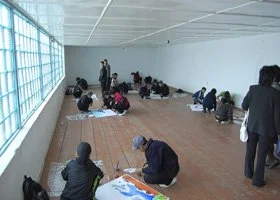 People sitting on the floor along the wall of a large room with a wooden floor, some working on art projects, others standing or walking.