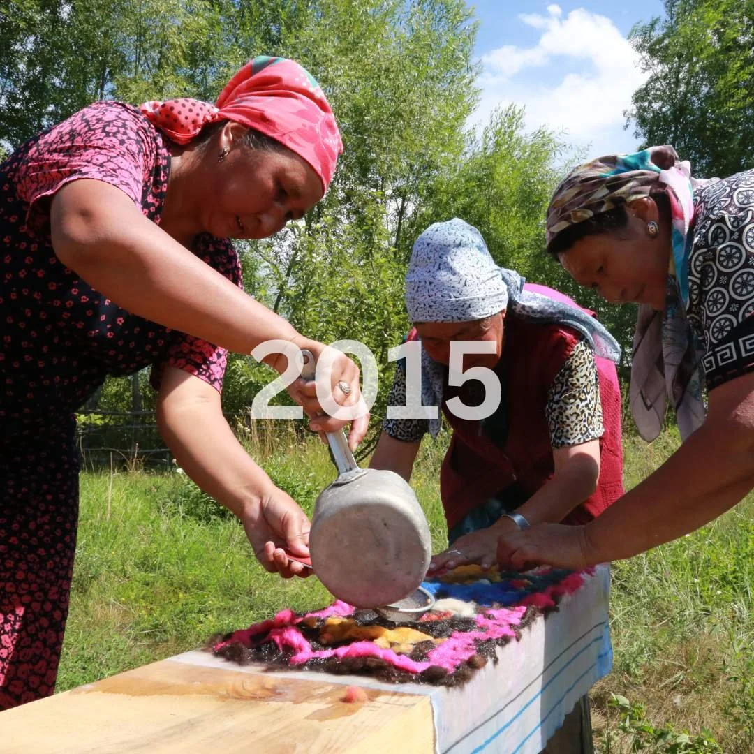Three women outdoors in sunny weather look down at a table with colorful felt and wool, working together on a craft project, with trees and blue sky in the background.