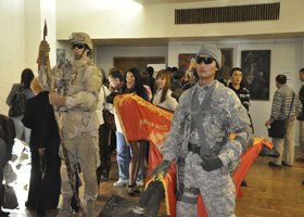 Two soldiers in camouflage uniforms and sunglasses carrying orange flags through a crowded indoor space with several people in the background.