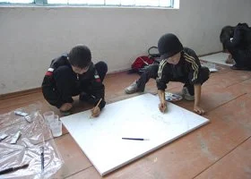 Two young men drawing on a large whiteboard on the floor.