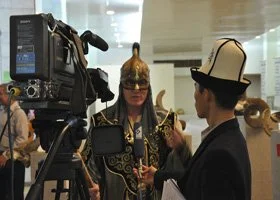 A woman being interviewed by a man wearing a hat, with a camera recording the scene in an indoor setting.