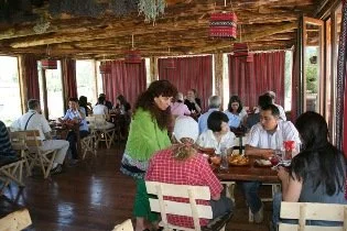 A busy restaurant with patrons dining at tables, and a waitress taking an order from a group of customers, in a rustic wooden interior with large windows.