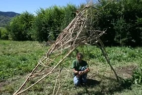 Person kneeling next to a large makeshift structure made of sticks and branches outdoors.