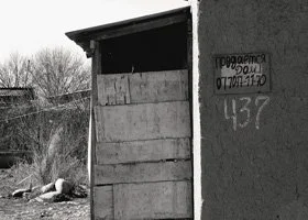 Close-up of a building corner with a window, sign, and graffiti that includes a date and number, in a rural area with trees and trash nearby.