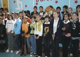 Group of children and a woman standing together indoors, possibly at a school event or celebration.