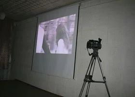 A projection screen displaying an image of two individuals in an outdoor setting. A video camera on a tripod is set up in front of the screen in a room with plain walls.