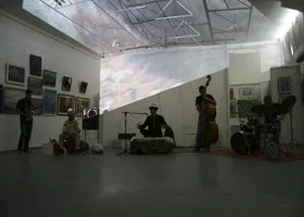 People performing music in an art gallery with artwork on the walls and a large projection on the back wall.