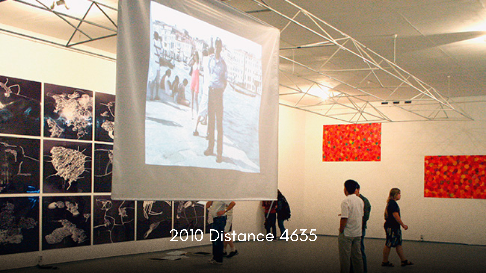 People walking around an art gallery with colorful abstract paintings on the walls and film projections on screens, including a large projected photo of a man and children on a beach. The caption reads '2010 Distance 4635'.