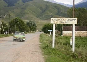 A rural road with a car driving on it, surrounded by green hills and a sign in a foreign script
