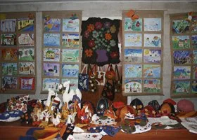 Display of colorful handmade crafts and artwork, including embroidered pictures, dolls, and decorative items on a table and wall.