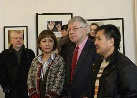 A group of five people standing indoors, engaged in conversation, with framed artwork on the wall behind them.