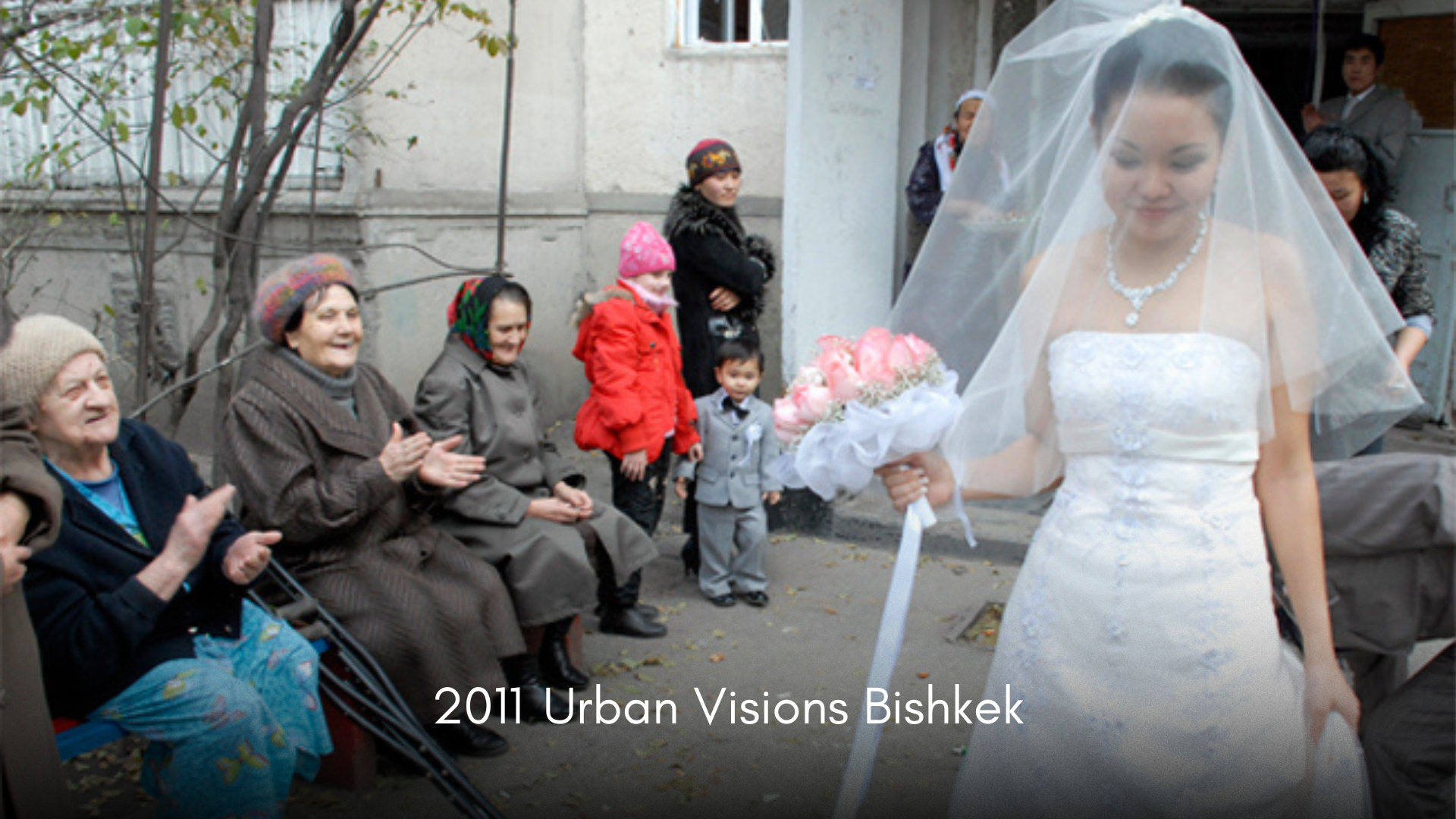 Bride in a white wedding dress holding a bouquet of pink roses, standing outdoors in front of seated elderly women and children, during a wedding celebration.