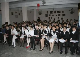 A large group of students dressed in formal attire sitting in a classroom or assembly hall, likely attending a school event or ceremony.