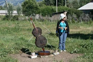 A woman wearing a cowboy hat, blue shirt, and jeans stands in a grassy field next to a guitar-shaped scarecrow with a watering can on top. There are bowls and containers on the ground nearby and a fence with trees in the background.