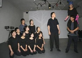 Group of people modeling in a dance studio, some standing and some sitting, dressed in black.