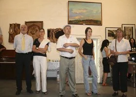 Group of six people standing in an art gallery, looking at artworks. One man holds a stack of papers, others have their hands by their sides or crossed, with paintings on the wall behind them.