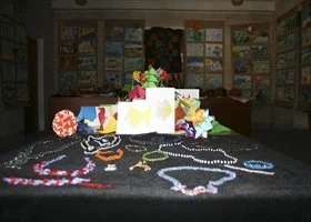 Collection of colorful handmade jewelry including necklaces and bracelets displayed on a dark surface with a backdrop of bookshelves and a decorated wall.