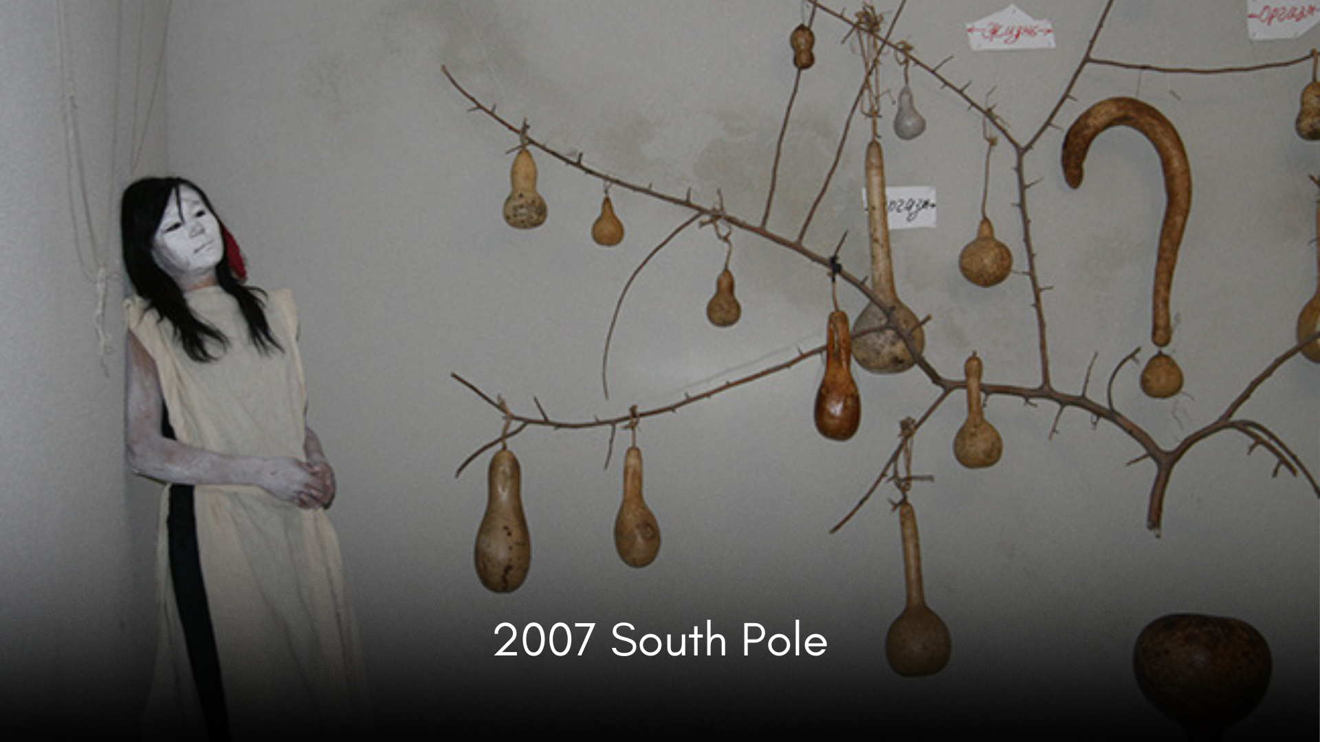 An art installation featuring a tree branch with gourd-like objects hanging from it. A person with face paint and dressed in white stands nearby, looking at the display.