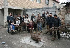 Group of people standing and sitting outdoors in an urban area near unfinished brick buildings, with some holding sticks and a tree branch on the ground.