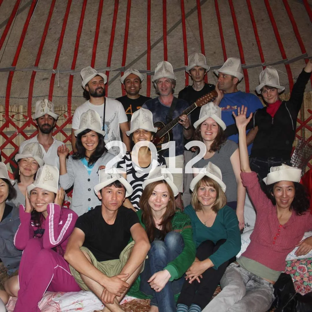 Group of people wearing white firefighter hats, some sitting and some standing, inside a yurt with red and beige walls. One person in the back is holding a guitar. The image has the year 2012 written across it.