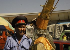A person dressed in traditional or costume attire, with a golden object on their head, possibly a headpiece or hat, while another person wearing a cap and uniform stands nearby. The scene appears to be outdoors with tents and vehicles in the backgrou
