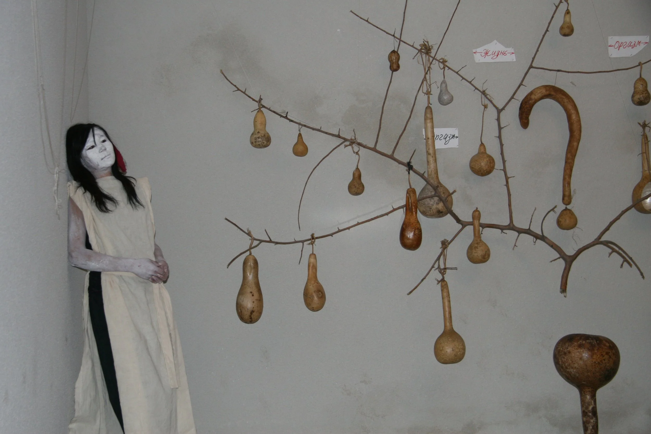 A mannequin with a painted white face and black hair stands beside a wall with a tree-like structure of branches hanging with various gourd-like objects and signs with writing in Cyrillic script.