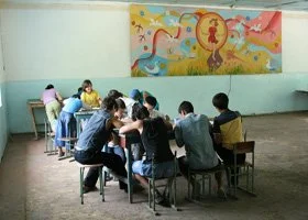 A group of children sitting around a table in a classroom, working on a project, with a colorful mural on the wall.