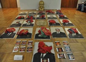 Display of framed photographs and artifacts on the floor, with some red cloths and a small golden bust in the center against a wooden wall.