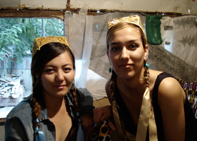 Two young women wearing traditional dresses and braids, standing inside a tent with a window and colorful textiles hanging in the background.