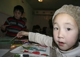Child wearing a gray beanie pointing at a colorful drawing on a table, with other children in the background.