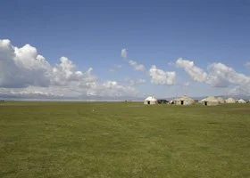 Open grassy field with a few small buildings in the distance under a partly cloudy blue sky.