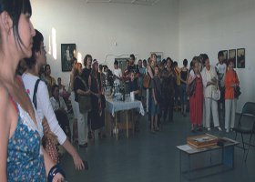 People gathered in an art gallery, standing and viewing artwork on the walls, with a table of items in the center of the room.