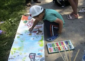 Child drawing on a large sheet of paper with colorful markers outdoors, with people and greenery in the background.