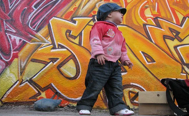 A young girl in a pink jacket and dark jeans stands in front of colorful graffiti on a wall, wearing a navy cap and white shoes, with a backpack and a cardboard box nearby.