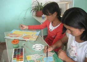 Two young girls painting and coloring at a table with art supplies in a classroom.
