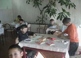 Children sitting at tables in a classroom, reading books and engaging in activities.