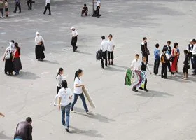 Group of people walking and standing in an open, paved area, some wearing traditional attire.