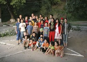 Group of children and adults posing outdoors in a parking lot with trees in the background.