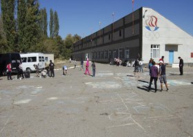 People walking around in a parking lot outside a large, rectangular building with multiple windows and a logo on the corner.