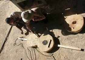 Two people working outdoors on a dirt surface, creating music with two homemade guitar-like instruments connected to microphones and cables.