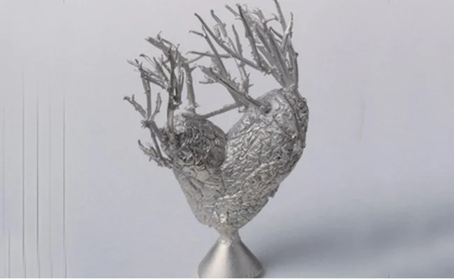 A silver sculpture with a heart shape and tree branches extending from the top, set against a plain white background.