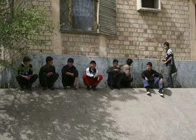 Seven boys sitting on the sidewalk against a brick building, with one standing and talking to them.