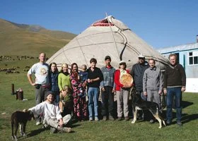 Group of people standing outside a traditional round yurt with green hills in the background.