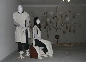 Two women wearing white masks in a dimly lit room with hanging objects and artifacts on the wall.