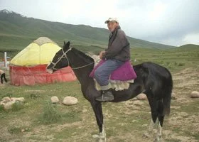 Person riding a black horse in a grassy landscape with yellow tents in the background